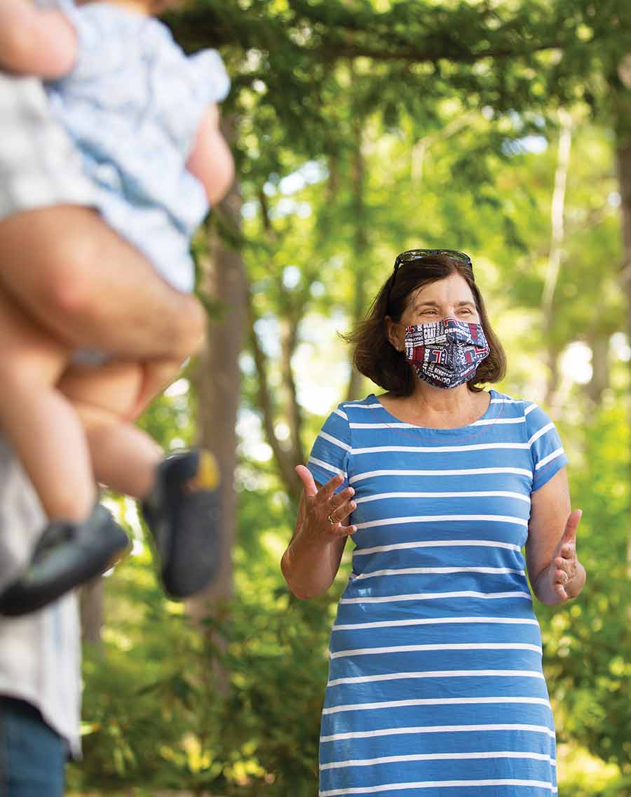Cinde in blue striped summer dress and face mask speaking in the park to a man with a young boy in his arms